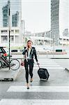 Woman walking outdoors, pulling wheeled suitcase, talking on smartphone