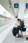 Woman sitting on train platform, using smartphone, suitcase beside her