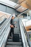 Woman on escalator, holding wheeled suitcase and smartphone, low angle view