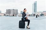 Portrait of businesswoman outdoors, sitting on suitcase, smiling
