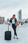 Portrait of businesswoman outdoors, holding wheeled suitcase, looking at smartphone