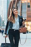 Businesswoman walking outdoors, pulling wheeled suitcase, holding smartphone and handbag, smiling