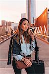 Young woman standing outdoors, using smartphone, wheeled suitcase beside her