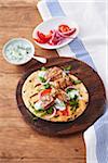 Pork souvlaki on a grilled pita on a wooden cutting board with side dishes of tzatziki and chopped tomatoes and sliced onions