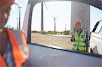 Smiling engineer using walkie-talkie at truck at sunny wind turbine power plant