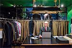 Senior tailor at desk using laptop in traditional tailors shop, portrait