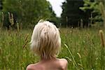 Bare shouldered blond haired boy in looking out at forest from long grass, rear view