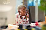 Businesswoman talking on smart phone, working at computer in office