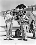 1940s TWO US NAVY PILOTS WEARING LEATHER FLYING HELMETS AND PARACHUTES STANDING READY BY PROPELLER BIPLANE