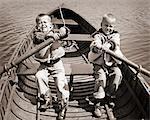 1950s 1960s PAIR OF BOYS SITTING IN BOAT EACH HOLDING ONE OAR STRUGGLING TO ROW