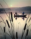 1980s TWO ANONYMOUS SILHOUETTED FISHERMEN SITTING IN MOTOR BOAT FISHING ON LAKE CATTAILS IN FOREGROUND