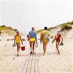 1970s FAMILY MOTHER FATHER TWO BOYS WALKING ON TO BEACH CARRYING CHAIRS UMBRELLA GEAR