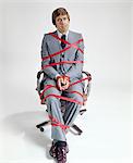 1970s BUSINESS MAN TIED UP IN RED TAPE SITTING ON OFFICE DESK CHAIR LOOKING AT CAMERA