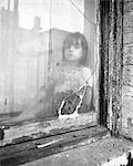 1970s POOR LITTLE GIRL LOOKING AT CAMERA OUT OF BROKEN WINDOW OF SLUM APARTMENT
