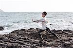 Young woman with brown hair and dreadlocks wearing white blouse standing on rocky shore by ocean, doing Tai Chi.