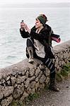 Young woman with brown hair and dreadlocks wearing headscarf sitting on dry-stone wall near ocean, taking picture with her mobile phone.