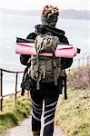 Rear view of young woman with brown hair and dreadlocks carrying backpack, walking along coastal hiking path.