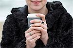 Close up of young woman wearing black furry jacket holding paper cup with hot drink.