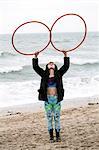 Young woman with brown hair and dreadlocks standing on a sandy beach by the ocean, balancing two hula hoops.