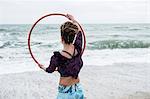 Young woman with brown hair and dreadlocks standing on a sandy beach by the ocean, balancing hula hoop.