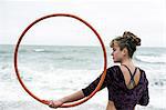 Young woman with brown hair and dreadlocks standing on a sandy beach by the ocean, balancing hula hoop.