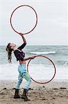 Young woman with brown hair and dreadlocks standing on a sandy beach by the ocean, balancing two hula hoops.