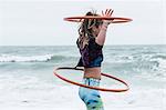 Young woman with brown hair and dreadlocks standing by the ocean, balancing two hula hoops.