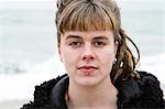 Portrait of young woman with brown hair and dreadlocks and a lip piercing wearing black furry jacket, looking at camera.