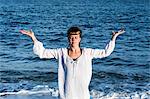 Young woman with brown hair wearing white blouse standing on a beach by the ocean, doing Tai Chi.