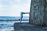 Young woman doing handstand by the ocean, with her legs against a wall.