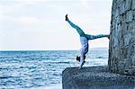 Young woman doing handstand on a wall by the ocean, one leg raised.