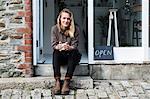 Smiling female owner of plant shop sitting on steps outside her store, looking at camera.