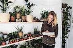 Female owner of plant shop standing next to a selection of plants on wooden shelves, holding digital tablet.