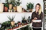 Female owner of plant shop standing next to a selection of plants on wooden shelves, holding digital tablet, smiling at camera.