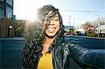 Portrait of young smiling woman with long curly black hair standing on street, looking at camera.