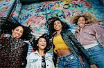 Low angle view of four young women with curly hair standing in front of shutter covered in colourful graffiti, smiling at camera.