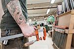 Closeup of an arm with tattoos and a hand holding a bar clamp in a large woodworking factory.