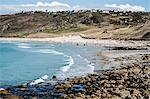 View along a coastline and sandy beach, waves breaking and people on the shore, with houses on the overlooking cliffs.