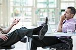 Two businessmen talking at a desk with one person's feet resting on top of the table