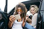 Portrait of two smiling young women with blond and brown curly hair sitting in car, taking selfie with mobile phone.