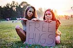 Young women sitting on grass holding free hugs sign at Holi Festival, portrait