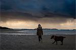 Young boy walking on beach with pet dog, looking at dramatic stormy sky, rear view, Lahinch, Clare, Ireland