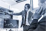 Businessmen trading stocks online. Stock brokers looking at graphs, indexes and numbers on multiple computer screens. Colleagues in discussion in traders office. Blue toned black and white.