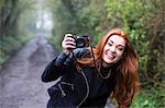 Smiling young woman with long red hair walking along forest path, taking pictures with vintage camera.