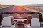 View from behind of senior couple in a convertible sports car driving on a highway at sunset in eastern Washington State, USA.