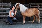 Smiling woman standing outside stable, holding a Shetland pony by its rein.