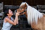 Smiling woman standing outside stable, holding a Shetland pony with a long blonde mane by its bridle.