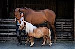 Woman standing outside stable, holding brown horse and a Shetland pony by their bridles.