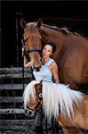 Woman standing outside a stable, holding a brown bay horse and a small Shetland pony by their reins.