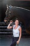 Smiling woman standing outdoor at stable, holding brown horse by rein.